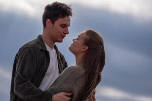 un joven Pareja abrazos, intercambiando oferta mira en contra un nublado playa fondo. el escena evoca romance y conexión en naturaleza, un fecha afuera. foto