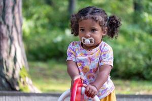 A thoughtful child with a pacifier, riding a toy in the park, captures a carefree summer moment, echoing the innocence of early childhood, and smartness from a young age. photo