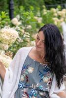 Adorned in a floral dress, a woman smiles gently amongst hydrangeas, her demeanor reflective of the wisdom and beauty found in life's natural cycles. photo