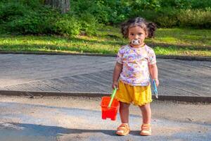 A curious toddler holds a red bucket and spade, pacifier in place, ready for sandbox adventures, early child development and curiosity about life. photo