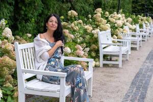 A pensive woman in a floral dress lounges on a white garden bench, her shawl draped casually, amidst a pathway of lush hydrangeas, evoking a moment of peaceful solitude. photo