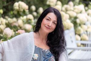 A smiling woman with black hair, wearing a floral dress and a white shawl, sits on a park bench surrounded by hydrangea bushes, radiating tranquility and contentment. photo