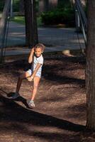 Young girl smiling on swing, dappled sunlight, park setting, wearing blue shorts and sandals, touching her hair. photo