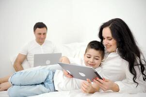 latest technology gadgets mom and son playing on tablet father sitting on white bed working on laptop remote work freelancer vacation family combine work and spend time with family together photo