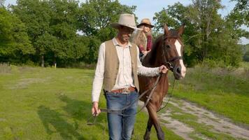 een jong cowboy is leidend een paard Aan welke zijn dochter is zitten. gelukkig cowboy familie. langzaam beweging. video