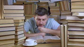 A bearded man reads a book in the library. Stacks of books and cup of tea on table. Concept of Reading and Education. video