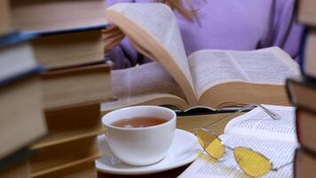 vrouw hand- flippen door de Pagina's van een boek Aan tafel in bibliotheek. stapels van boeken, een kop van thee en bril in de voorgrond. concept van lezing en onderwijs. dichtbij omhoog video