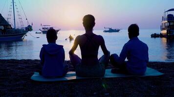 famiglia fare yoga. mamma con bambini siamo Meditare su il mare spiaggia a tramonto. contento e salutare famiglia. video