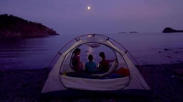 Mama mit Kinder in der Nähe von das Zelt beim Nacht. Sie sitzen auf das Strand von das Mittelmeer Meer. video