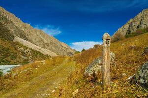 A rustic wooden milepost stands by a meandering path through a golden valley framed by imposing grey mountains, under a vast blue sky with scattered clouds, evoking tranquility and remoteness. photo