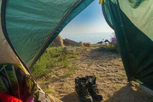 View of the coastal landscape from inside the tent with sleeping bags and a backpack. Hiking boots. Sea merges with a blue sky. To the right of the tent, stones and other camping items are arranged photo