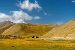 Vast golden grasslands stretch beneath rolling hills, leading to the ancient Tash Rabat caravanserai, visible in the distance, a serene, remote landscape under a vast blue sky with white clouds photo