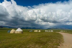 A serene nomadic settlement of circular yurts unfolds along the verdant plains of Son-Kul, embraced by the grandeur of distant mountain silhouettes and a canvas of sprawling, cloud-laced blue skies. photo