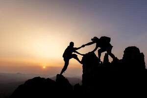 Silhouette of person hikers climbing up mountain cliff and one of them giving helping hand. People helping and, team work concept. photo