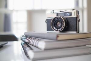 A vintage film camera sits on a table in an office and a book stands out in the background. A vintage film camera with a simple design sits on a table in an office and has copy space for text. photo