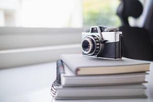 A vintage film camera sits on a table in an office and a book stands out in the background. A vintage film camera with a simple design sits on a table in an office and has copy space for text. photo