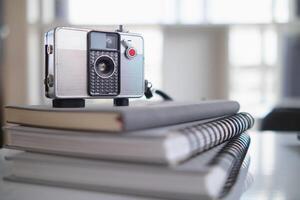 A vintage film camera sits on a table in an office and a book stands out in the background. A vintage film camera with a simple design sits on a table in an office and has copy space for text. photo