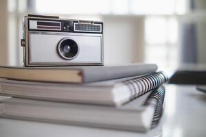 A vintage film camera sits on a table in an office and a book stands out in the background. A vintage film camera with a simple design sits on a table in an office and has copy space for text. photo