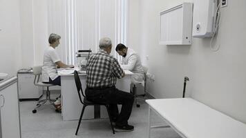 Senior man patient sitting on chair by the table of a doctor and a nurse. Clip. Medical checkup visit at the hospital. photo