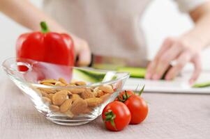 de cerca en el mesa vaso plato con nueces Almendras Tomates y campana pimientos en antecedentes hembra manos sabroso Fresco vegetales ensalada foto para antecedentes Cocinando borroso cristalería sitio para tu anuncio