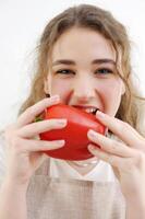 gracioso alegre adolescente niña joven mujer mordiendo rojo pimienta sano comiendo delicioso comida Cocinando ensalada atentamente astuto Mira sonrisa hermosa dientes marrón pelo delantal cocina de cerca delicado manicura salud foto