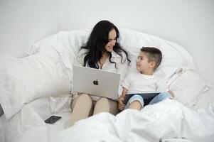 mom and son sit in a white bed in their hands Apple technology laptop MacBook tablet iPad and mobile phone iPhone 13 pro Max look at each other smile uses the latest technology love care wealth is photo