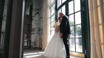 Bride and Groom Posing by Sunlit Window. The bride holds a bouquet of roses and dried flowers, while the groom stands close, both sharing a tender moment. video