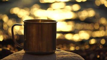 Close-up of a travel stainless mug standing on a rock. Small waves on golden warm water surface with bokeh lights from sun. A concept of the world of beauty, nature and outdoor travel. Selective Focus video