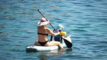 Sea woman sup. A happy positive woman in hat with family relaxing in sea, aerial back view of family on SUP board floating on calm water. Active lifestyle at sea. Summer vacation. Slow motion video