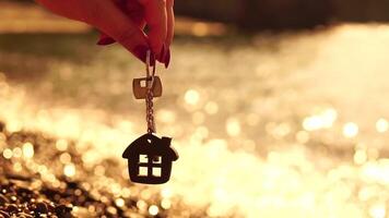 Keys with a house-shaped keychain in a woman's hand on sea beach background. The concept of buying a home, apartment, real estate, young family, home loan, rental housing. Close-up, Slow motion video