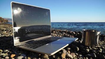 Silhouette of laptop and cup on the beach over sea sunset. Sparkling sea water on background. Freelancer, blogger working on notebook on the beach. video