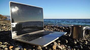 Silhouette of laptop and cup on the beach over sea sunset. Sparkling sea water on background. Freelancer, blogger working on notebook on the beach. video