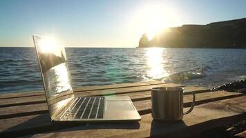 Silhouette of laptop and cup on the beach over sea sunset. Sparkling sea water on background. Freelancer, blogger working on notebook on the beach. video