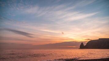 Sunset Sky Clouds Timelapse. A red burning sunset over the sea rocky volcanic cliff. Abstract nature summer ocean sea background. Small waves on golden warm water surface with bokeh lights from sun. video