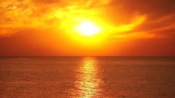 A red burning sunset over sea with rocky volcanic cliff. Abstract nature summer ocean sea background. Small waves on golden warm water surface with bokeh lights from sun. Weather and climate change video
