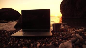 Silhouette of laptop and cup on the beach over sea sunset. Sparkling sea water on background. Freelancer, blogger working on notebook on the beach. video