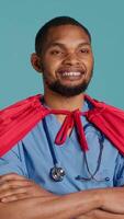 Vertical Smiling male nurse portraying superhero wearing red cape, isolated over studio background. Portrait of cheerful heroic medic posing as hero in costume with mantle, camera A video