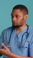 Vertical Portrait of african american nurse pointing hands towards empty space, doing advertising, talking with audience. Healthcare employee doing promotional ad, showing copy text, studio backdrop, camera A video