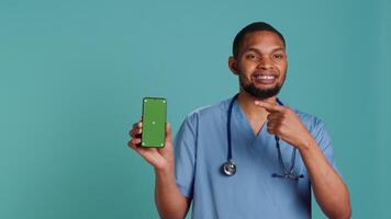 retrato do alegre masculino enfermeira apontando dedo para verde tela Smartphone. otimista hospital empregado segurando croma chave telefone, isolado sobre azul estúdio fundo, Câmera uma video