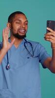 Vertical Male nurse waving hand in videocall meeting using phone, saluting patient. Caretaker chatting with patient in online telemedicine call using cellphone, studio backdrop, camera B video