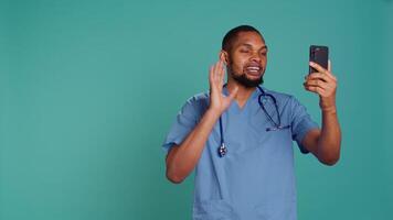 Male nurse waving hand in videocall meeting using phone, saluting patient. Caretaker chatting with patient in online telemedicine call using cellphone, studio backdrop, camera B video