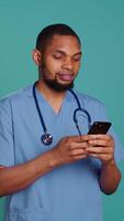 Vertical Male nurse using mobile phone to scroll on social media feed during hospital job shift break. Healthcare worker using smartphone to text friends, studio backdrop, camera B video