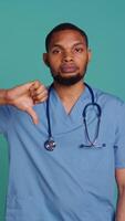 Vertical Upset nurse showing thumbs down signs while at work, isolated over blue studio background. Disappointed healthcare professional doing frenetic disapproval gestures, camera B video