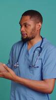 Vertical Portrait of african american nurse pointing hands towards empty space, doing advertising, talking with audience. Healthcare employee doing promotional ad, showing copy text, studio backdrop, camera B video