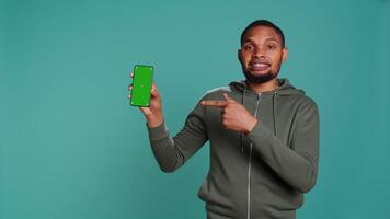 Portrait of african american man doing influencer marketing using green screen phone, studio background. BIPOC person holding empty copy space mockup smartphone device, camera B video