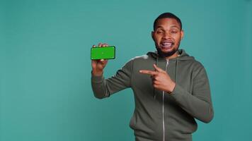Portrait of upbeat content creator doing influencer marketing using green screen mobile phone. Cheerful person holding chroma key cellphone device, isolated over studio backdrop, camera B video