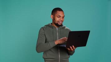 Smiling man scrolling on laptop, using internet and credit card to pay for products. African american person happily browsing online shopping websites, isolated over studio background, camera B video