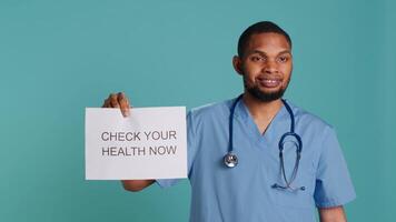 Portrait of upbeat nurse smiling, holding sign urging people to do doctor appointments, studio background. African american healthcare worker talks about importance of preventive healthcare, camera A video