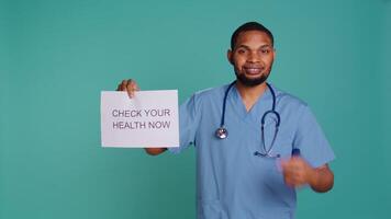 Portrait of upbeat nurse smiling, holding sign urging people to do doctor appointments, studio background. African american healthcare worker talks about importance of preventive healthcare, camera B video