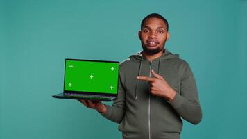 Smiling man holding isolated screen laptop, doing recommendation, talking with audience. Cheerful person pointing towards chroma key notebook, studio background, camera B video
